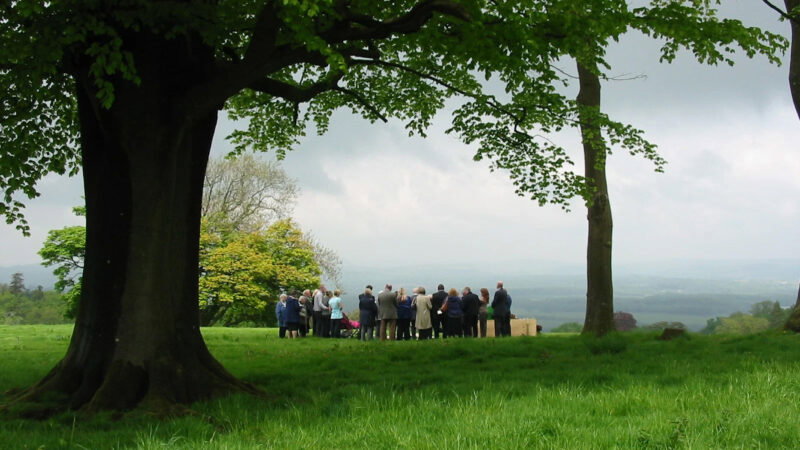 A group of people gathered outdoors in a grassy field for a natural burial, standing in a circle near large trees under a cloudy sky, with distant hills visible in the background—reflecting thoughtful funeral arrangements Cardiff.