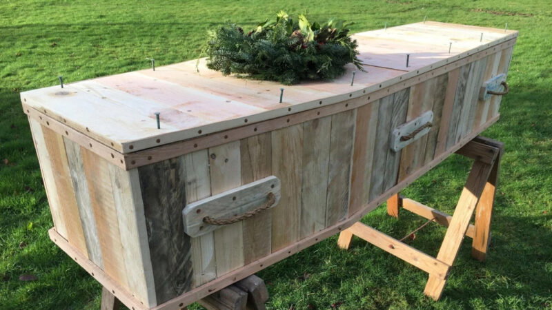 A wooden coffin with rope handles and visible nails sits on wooden trestles outdoors on green grass at Hundy Mundy, topped with a wreath of greenery—perfect for woodland burial arrangements.