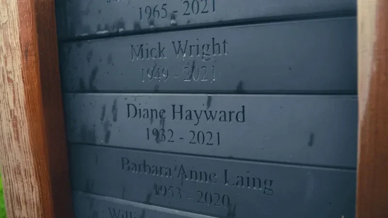 Close-up of a memorial plaque with engraved names and years, including Mick Wright (1949–2021) and Diane Hayward (1982–2021). The plaque, wet from raindrops, forms part of the thoughtful Hundy Mundy woodland burial arrangements.