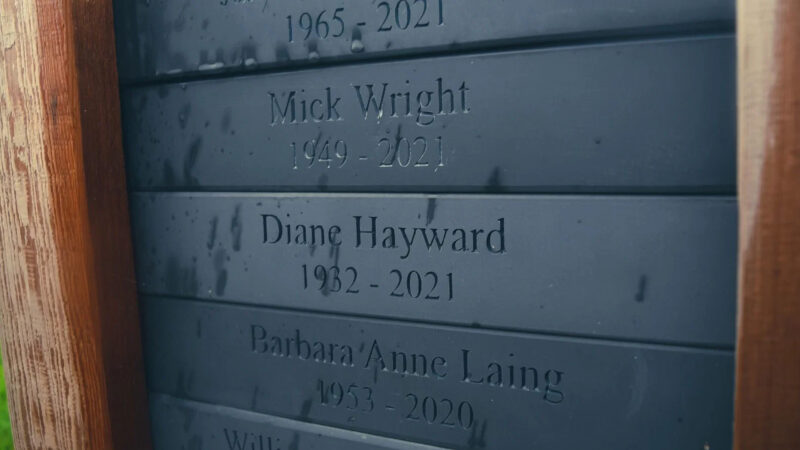 Close-up of a dark blue Hundy Mundy woodland burial memorial plaque with engraved names and years, including Mick Wright (1949–2021), Diane Hayward (1962–2021), and Barbara Anne Laing (1953–2020), water droplets on the surface.