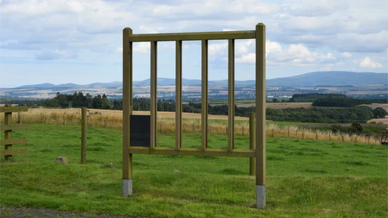 A large empty wooden sign frame stands in a grassy field, with rolling hills, farmland, and a cloudy sky in the background—an inviting scene reminiscent of the tranquil setting offered by Hundy Mundy woodland burial arrangements.
