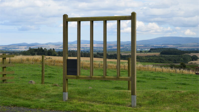 A large empty wooden sign frame stands in a grassy field with rolling hills, farmland, and a partly cloudy sky beyond—an inviting scene reminiscent of Hundy Mundy woodland burial memorials.