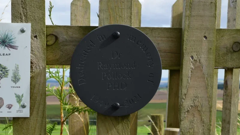 A round, black memorial plaque for Dr Raymond Pollock, PhD (1939–2021) is mounted on a wooden fence, next to a botanical illustration. The background shows a distant landscape and sky, evoking the serene atmosphere of Hundy Mundy woodland burial arrangements.