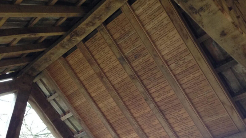 The ceiling of a wooden structure lined with many rectangular plaques, like those at Usk natural burial memorials, each engraved with text in neat rows along the beams. The plaques commemorate individuals or events as natural light filters through the structure.