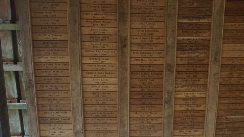 Weathered wooden ceiling panels at Usk natural burial memorials are engraved with numerous names and dates in columns, likely as a tribute or memorial. The rustic panels suggest an outdoor or nature-inspired structure.