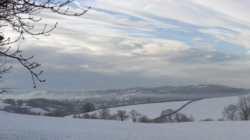 A snowy landscape blankets the Usk Natural Burial Meadow, with rolling hills and bare trees under a cloudy winter sky. Faint distant hills and fields are visible, snow covering the ground and branches in the peaceful foreground.