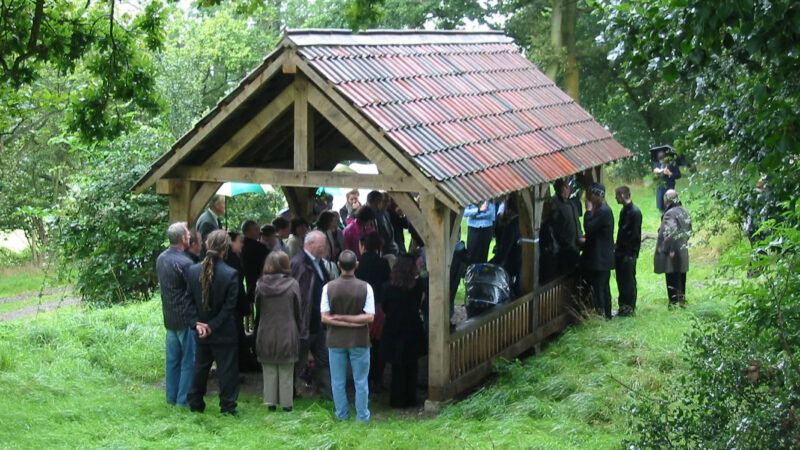A group of people gather under and around a wooden shelter with a tiled roof in the grassy, wooded Usk Natural Burial Meadow. Some attendees hold umbrellas, suggesting rain, as they attend a formal outdoor event.