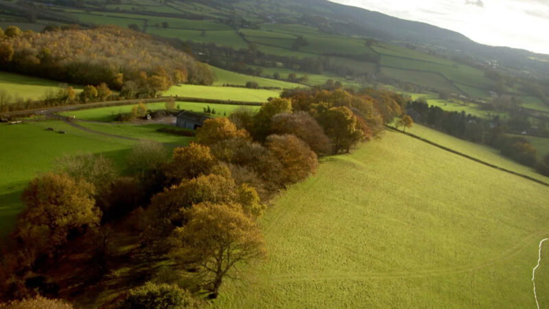 Aerial view of a rural landscape with green fields, autumn trees, a farmhouse, and rolling hills in the background under a partly cloudy sky, featuring the serene Usk Natural burial meadow nestled among nature’s beauty.