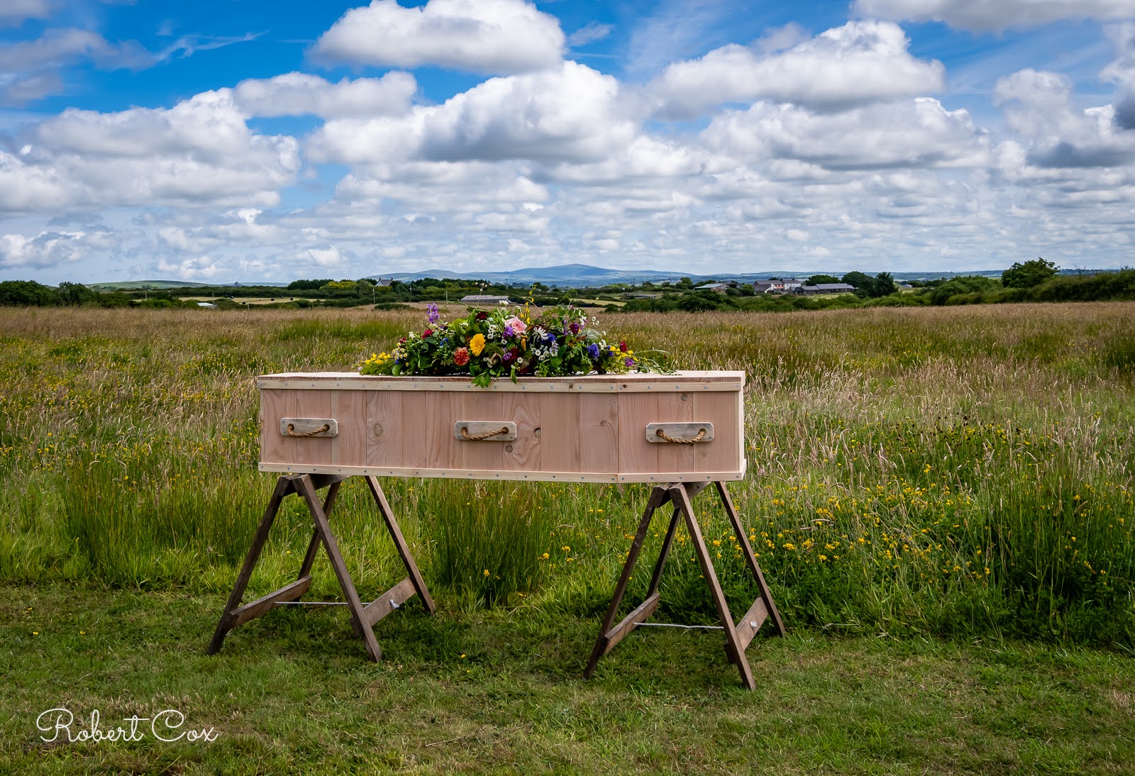 A wooden coffin decorated with colourful flowers rests on trestles in a grassy field under a partly cloudy sky at the peaceful Pembrokeshire Natural Burial Meadow.