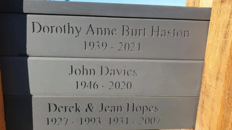 Three grey memorial plaques with engraved names and dates—Dorothy Anne Burt Haston (1939-2021), John Davies (1946-2020), and Derek & Jean Hopes (1927-1993, 1931-2007)—are mounted on a wooden structure, a quiet tribute often found in Arrangements Pembrokeshire Natural Burial Meadow.