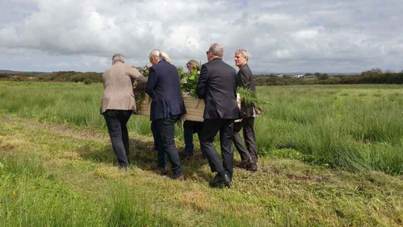 Six people in formal clothing carry a coffin adorned with greenery through a grassy field, likely part of Arrangements Pembrokeshire’s natural burial meadow, participating in an outdoor funeral procession under a cloudy sky.