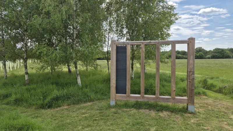 A wooden frame with a narrow black panel stands on grass near trees in a green field under a partly cloudy sky, evoking the tranquil setting typical of woodland burial arrangements in Henley-on-Thames.