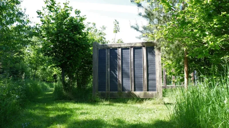 A solar panel is mounted vertically on a wooden frame surrounded by lush, green trees and grass in a sunlit outdoor setting, evoking the peaceful arrangements often found at a Henley-on-Thames woodland burial ground.