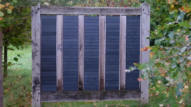 A wooden garden gate with vertical slats and a black panel in the centre stands on green grass, surrounded by leafy plants and trees—evoking the tranquil beauty of Arrangements Henley-on-Thames woodland burial settings.