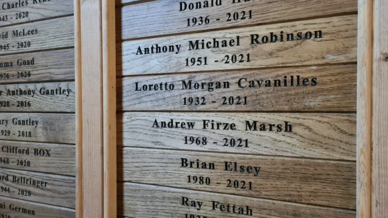 Wooden memorial plaques display engraved names and years of birth and death, including Anthony Michael Robinson (1951–2021), Loretto Morgan Cavanilles (1932–2021), and others, as part of thoughtful arrangements at Dorset Downs natural burial grounds.