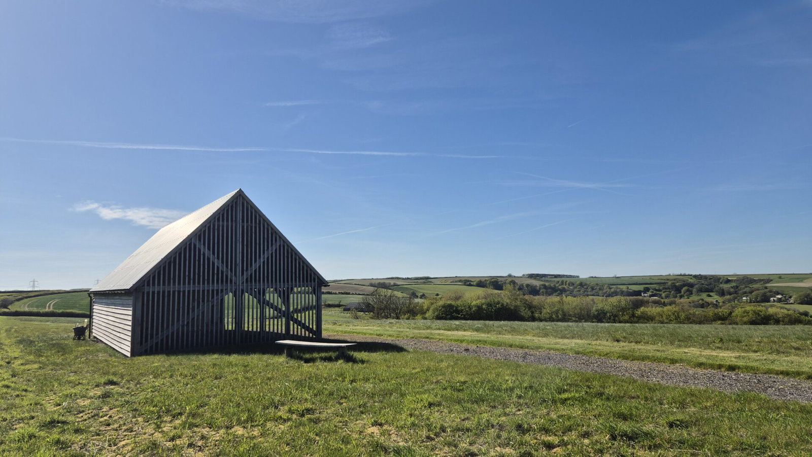 A wooden barn with a slanted roof stands on a grassy field under a clear blue sky, nestled among the rolling hills and scattered trees of the Dorset Downs, with a gravel path winding through this peaceful natural burial landscape.