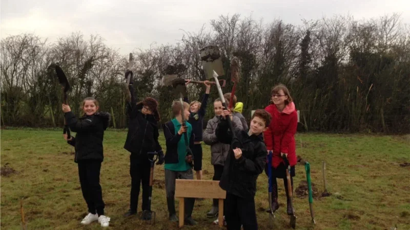 A group of children holding spades and trowels stand on grass in front of trees, smiling at the camera, some wearing jackets and wellies. A small wooden sign, part of the natural burial arrangements in Aylesbury Vale, is planted in the ground in front of them.