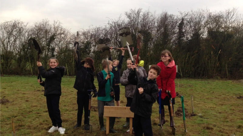 A group of children stand on grass holding spades and gardening tools, smiling at the camera near trees and bushes. They are dressed in jackets, enjoying nature close to the Aylesbury Vale natural burial memorials.