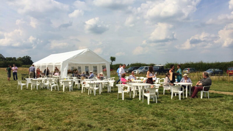 People are seated at white plastic tables and chairs on grass beside a white marquee during an outdoor event on a sunny day in Aylesbury Vale. Cars are parked in the background under a partly cloudy sky, adding to the peaceful arrangements.