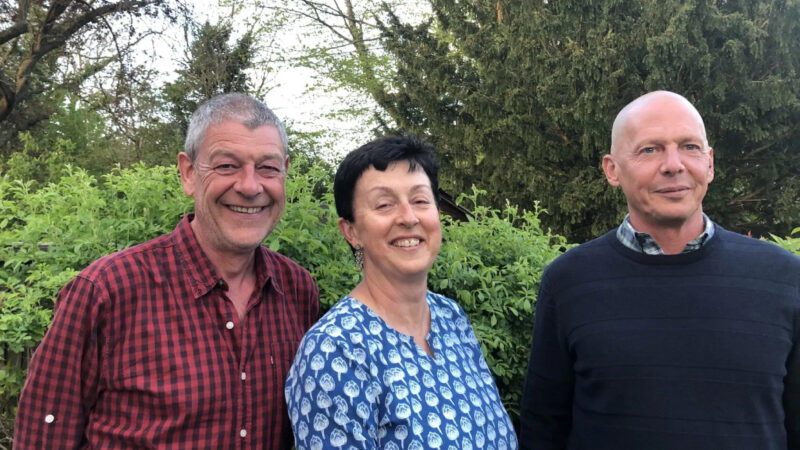Three adults stand outdoors in front of green bushes and trees, smiling at the camera. This warm moment captures the spirit of About Aylesbury Vale Natural Burial, with each person dressed in vibrant, comfortable attire.