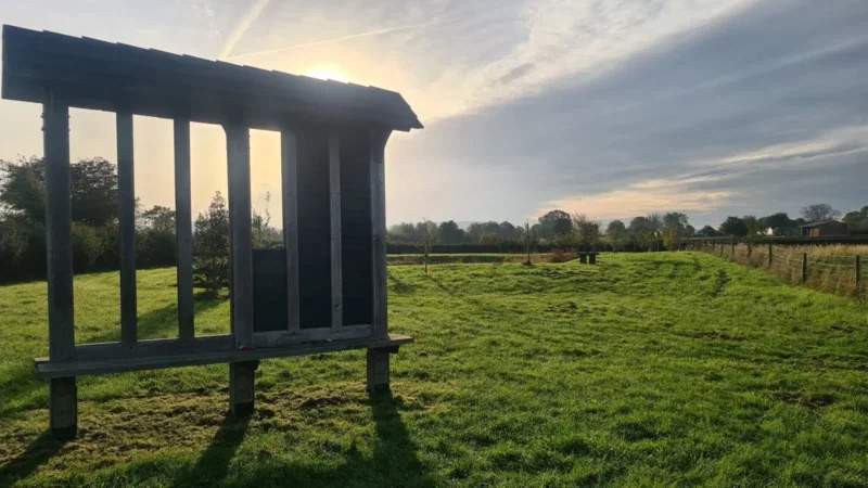 A wooden structure stands on a grassy field under a partly cloudy sky with sunlight streaming through. Trees and a fence line the background, creating a peaceful rural scene—an ideal setting for natural burial arrangements in Aylesbury Vale.