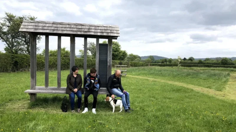 Three people sit on a wooden bench shelter in a grassy field, with a small dog standing beside the person on the right. The serene landscape of Aylesbury Vale unfolds around them—trees, hedges, and hills are visible in the background under a cloudy sky—suggesting tranquil moments perhaps spent reflecting or making arrangements for a natural burial.