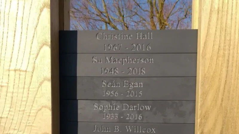 A memorial plaque lists names and years: Christine Hall 1967–2016, Su Macpherson 1948–2018, Seán Egan 1956–2015, Sophie Darlow 1933–2016, and John B. Willcox. Behind the tribute to these lives connected with natural burial arrangements in Aylesbury Vale, trees and blue sky are visible.