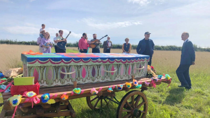 A group of people stand and play music around a colourful, decorated coffin on a wooden cart in Dorset Natural Burial Meadow. Bright paper flowers and fabric adorn the cart and coffin under a blue sky in the grassy field.