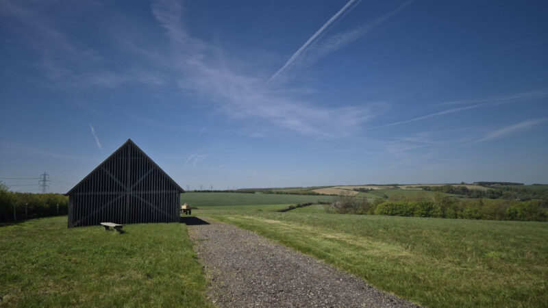 A small wooden barn stands beside a gravel path in a wide, green field at Dorset Natural Burial Meadow, under a blue sky with wispy clouds and vapour trails, with rolling hills and trees in the distance.
