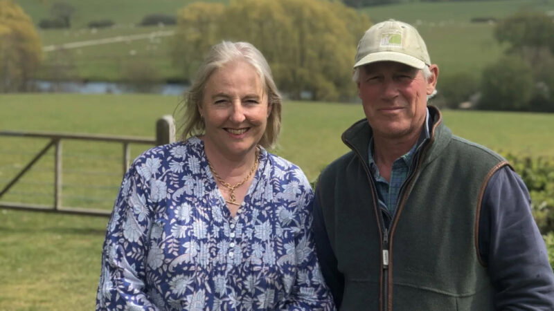 A smiling woman in a blue floral shirt stands next to a man in a green cap and waistcoat at Dorset Downs Natural Burial, outdoors in a grassy, rural area with trees, fields, and a wooden fence in the background.