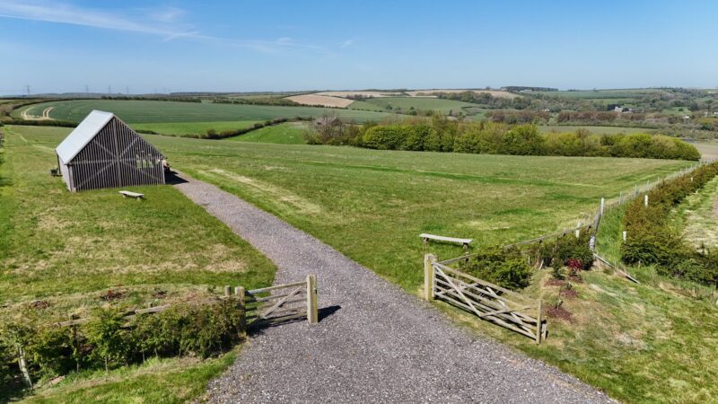 A gravel path leads through an open wooden gate towards a modern wooden shelter on a grassy hill at Dorset Natural Burial Meadow, surrounded by rolling green fields under a clear blue sky.