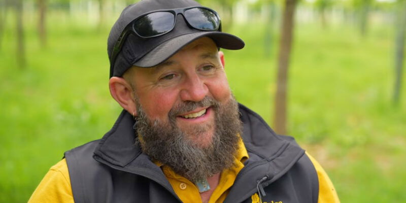 A bearded man wearing a black cap, sunglasses, and a black vest over a yellow shirt smiles while sitting outdoors in a green, grassy area with trees in the background, proudly sporting his Gravedigger of the Year hat.