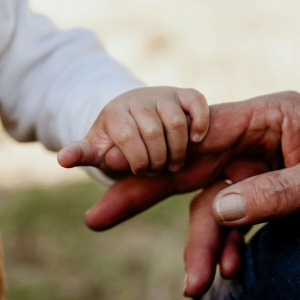 A close-up of a baby’s hand gently gripping an adult’s finger. The adult’s hand appears weathered, contrasting with the baby’s smooth skin. The background is softly blurred.