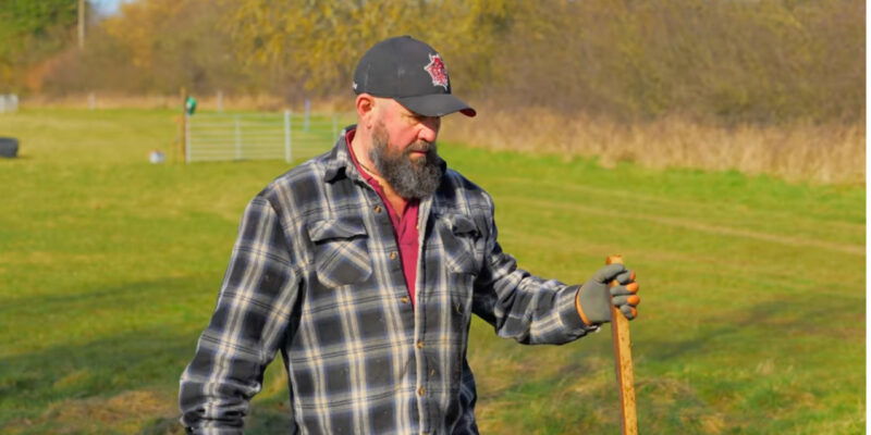 A bearded man, dressed in a plaid shirt, gloves, and a baseball cap, stands outdoors on a grassy field holding a wooden stick—an image befitting the Gravedigger of the Year amid trees and a distant gate.