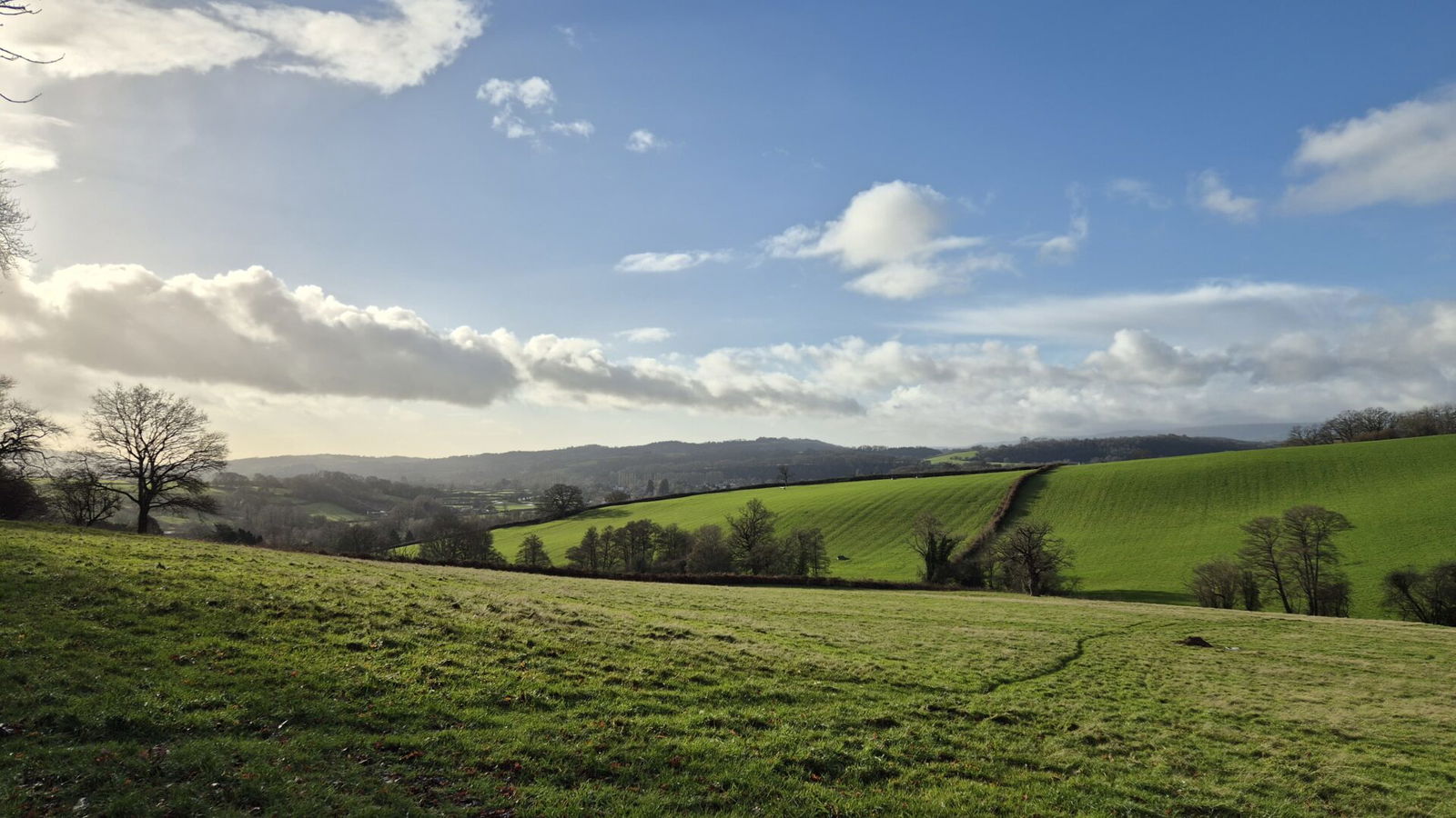 A scenic countryside landscape with rolling green hills, scattered trees, and a bright blue sky partly covered by white clouds. The sunlight casts soft shadows across the grassy fields.
