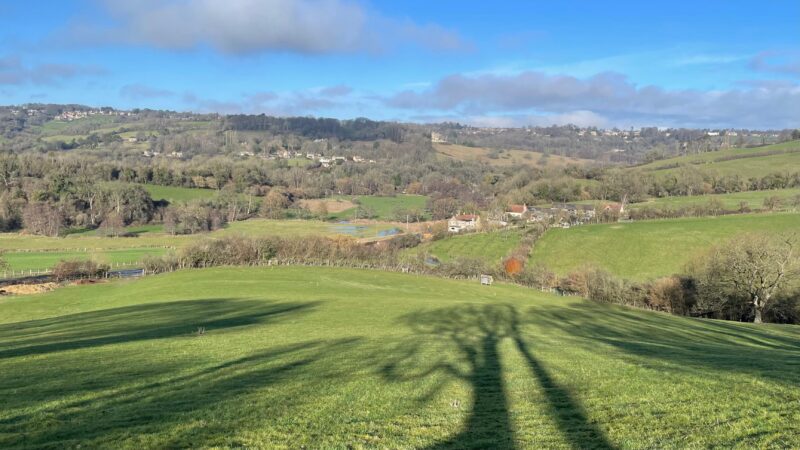 A sunlit rural landscape with rolling green hills, scattered trees, and distant houses. A large tree casts a long shadow across a grassy field under a mostly clear blue sky with a few clouds.