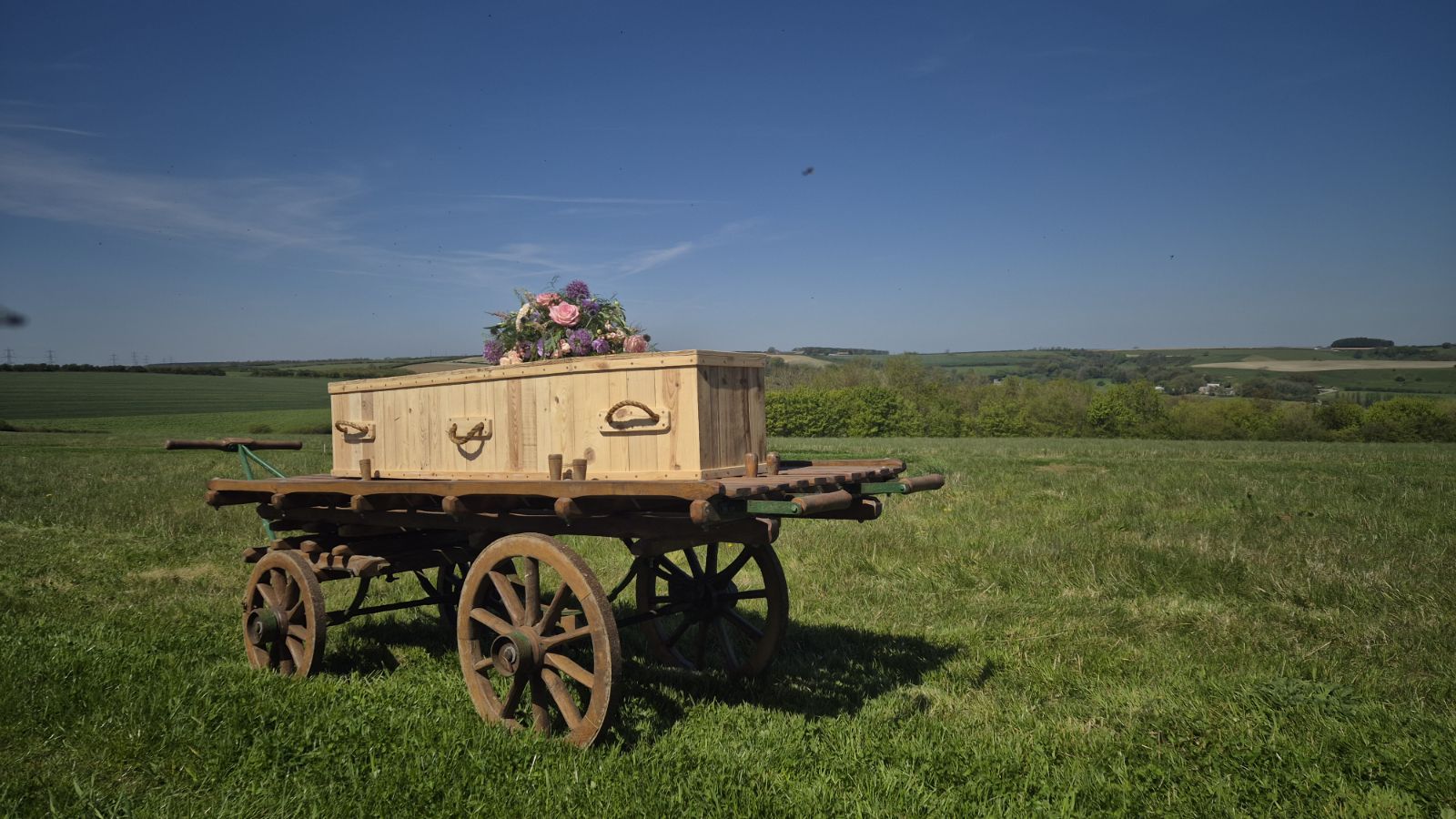 A wooden coffin adorned with flowers rests on an old wooden cart in a grassy field under a clear blue sky, with rolling green hills in the background.