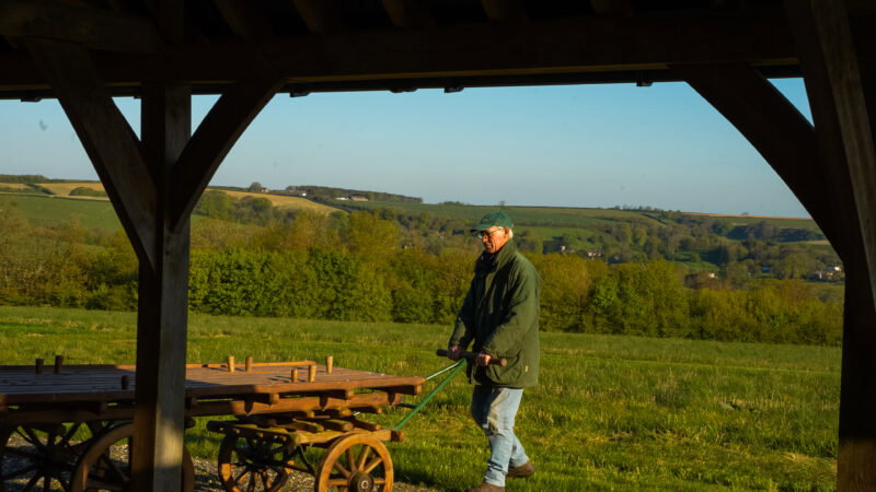 An older man in a green coat and cap pulls a wooden cart under a wooden shelter, with green fields and hills—where natural burial grounds have long grass—in the background on a sunny day.