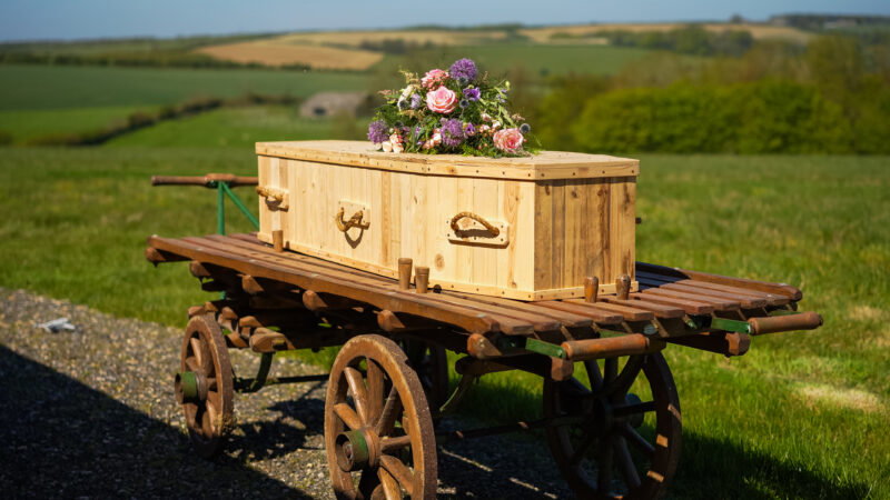A wooden coffin with flower arrangements rests on an old wooden trolley outdoors, surrounded by the long grass typical of natural burial grounds and distant rolling hills under a clear sky.