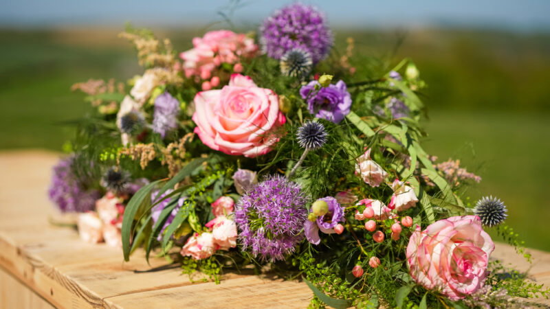 A colourful bouquet of pink roses, purple alliums, and green foliage rests on a wooden surface outdoors, echoing why natural burial grounds have long grass in their serene, untouched surroundings.