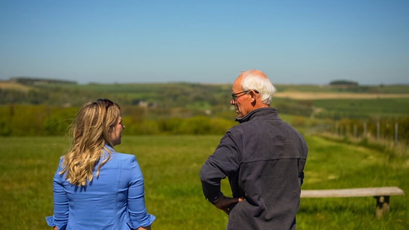 A woman and an older man stand outdoors in a grassy field, discussing why natural burial grounds have long grass. A bench sits nearby, with rolling hills and blue sky in the background.
