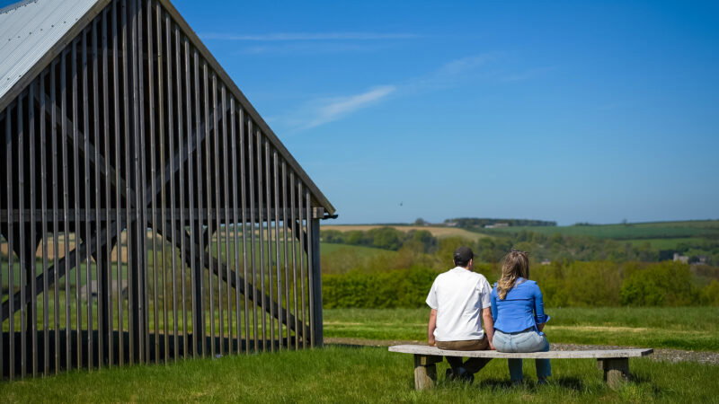 A man and woman sit on a bench in front of a wooden building, looking at a scenic landscape of green fields and blue sky, wondering why natural burial grounds have long grass.