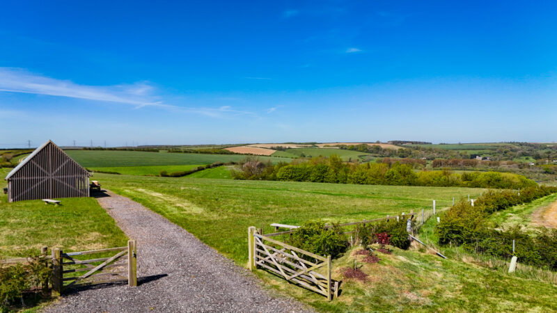 A wooden shed stands beside a gravel path and open gate, overlooking green fields dotted with long grass—much like those found in natural burial grounds, where the grass is left to grow freely to support biodiversity—and rolling hills beneath a clear blue sky on a sunny day.